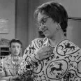  
Fotografia z filmu Čert nespí (1956). V pozadí za stolom sedí Ivan Krivosudský (účtovník Cyril Kvasnička), v popredí stojí Oľga Vronská (účtovníčka Pipulková)
